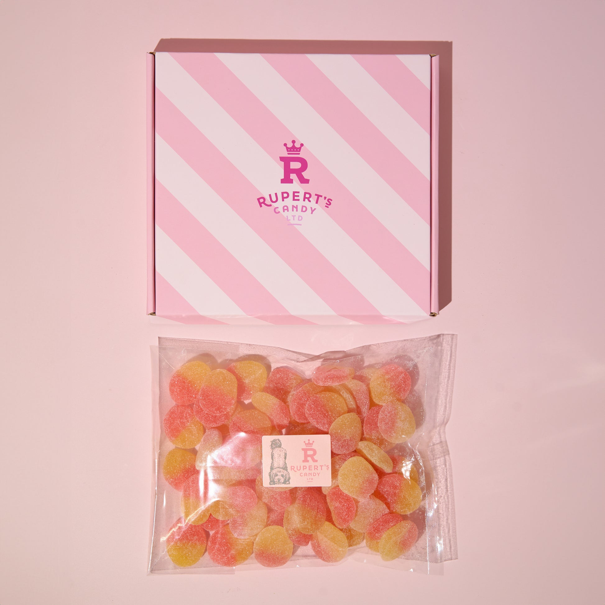 Image of our pink rupert's candy box with a bag of fizzy peaches laid underneath it- Rupert's candy